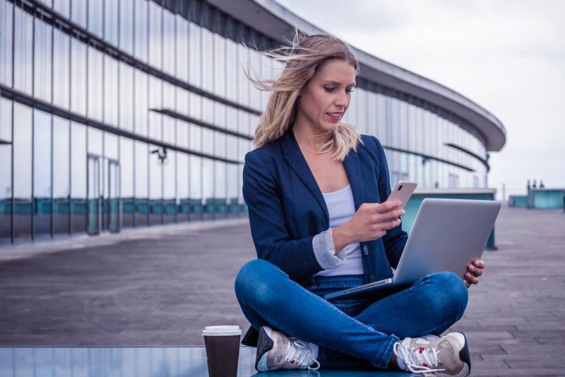 young woman, sitting outside, in front of a glass building, takes a picture with her mobile phone, on her laptop screen. The wind blows his blonde hair away.